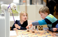 A young boy and girl play the Pikes and Ladders game in the main gallery. The girl is holding a white toy dog, Boye.